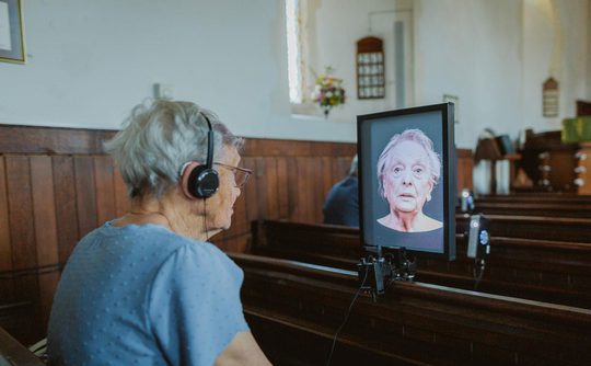 A person siting on a church pew, wearing head phones and watching a portrait video of a talking head