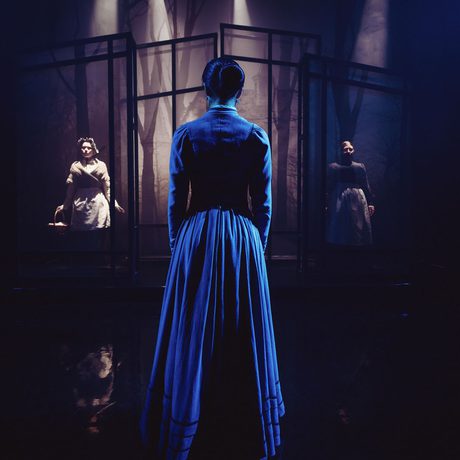 A person in a dress facing away to two performers behind lit screens.