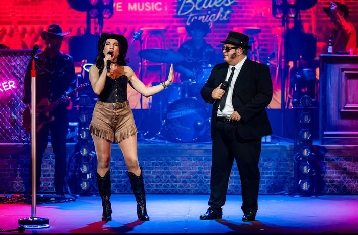 A live band on stage. One person is singer another person is dressed as a Blues Brother.