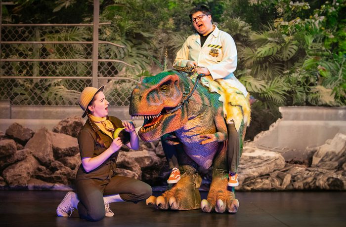 A person riding on the back of a dinosaur puppet and another person kneeling beside them.