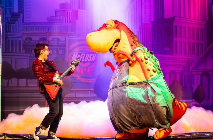 A person playing the guitar and a person in a large dinosaur costume dancing.
