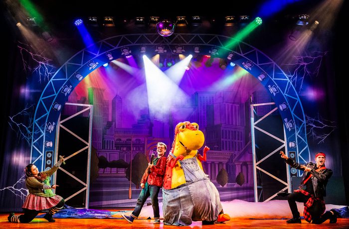 A person playing the guitar and a person in a large dinosaur costume on a stage.