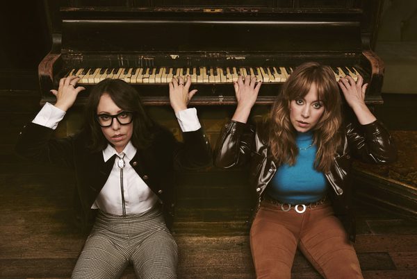 Flo and Joan sitting on the floor and playing a piano which is behind them.