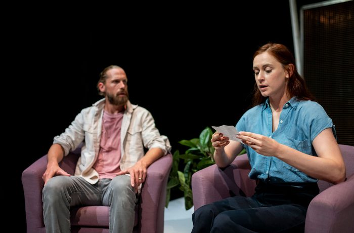 Two people sitting in armchairs. One person is reading a small note.