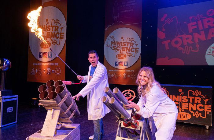 Two scientists conducting an experiment. One is holding a long pole with fire on the end.