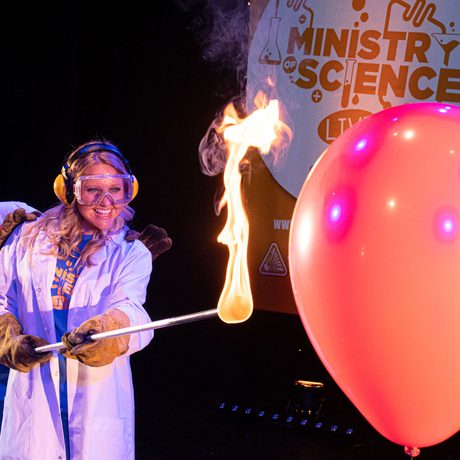 Two people in lab coats poking a balloon with a flaming stick.