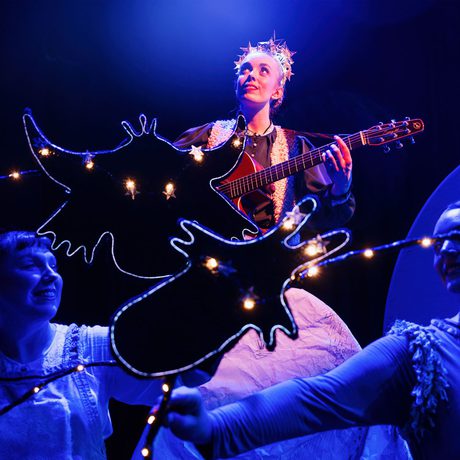 Three performers in dim lighting, one playing a guitar and the other two using light up puppets.