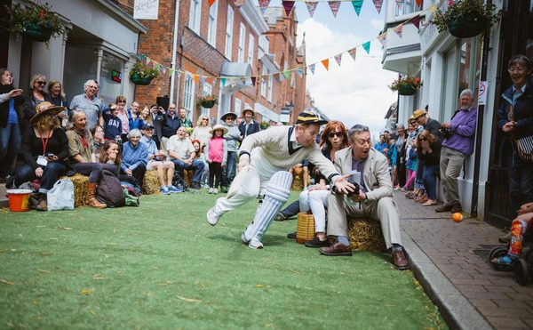 People sitting in the street. One person is dressed as a cricket player and catching a ball.
