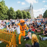 A person in an orange space suit running through a large crowd. Winchester Cathedral is in the background.