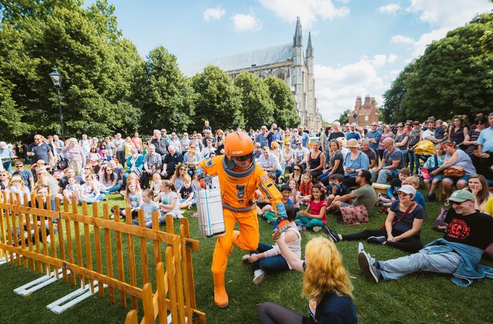 A person in an orange space suit running through a large crowd. Winchester Cathedral is in the background.