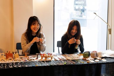 Two people smiling an laughing whilst sitting behind a stall selling jewellery
