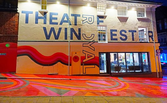 A nighttime photo of Theatre Royal Winchester, with colourful artwork across the building and in the streets