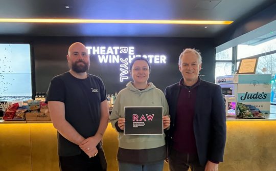 Three people standing together, holding a sign that says "RAW"