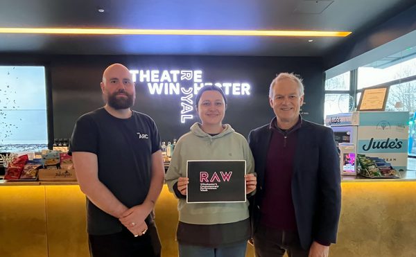 Three people standing together, holding a sign that says "RAW"
