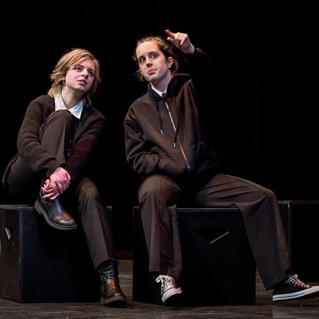 Two young performers on a stage, dressed in black and sitting on boxes