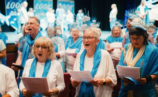 A group of people dressed in white and blue singing from song sheets.