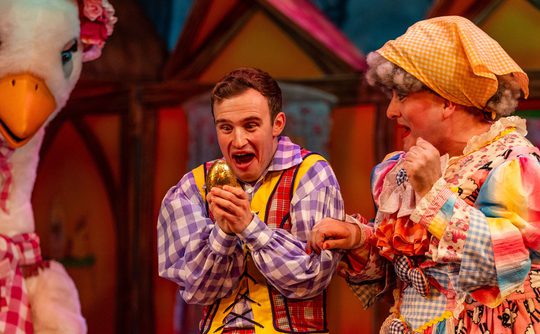 A panto performer looking excited and holding a golden egg, accompanied by a Goose and the panto Dame.