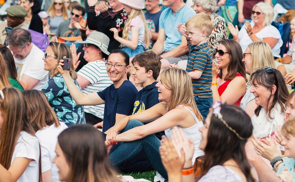 A large group of people sitting a grass lawn, laughing and clapping.