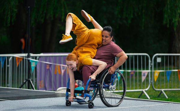 Two people riding a wheelchair on a stage. One person is seated and the other is doing a handstand.