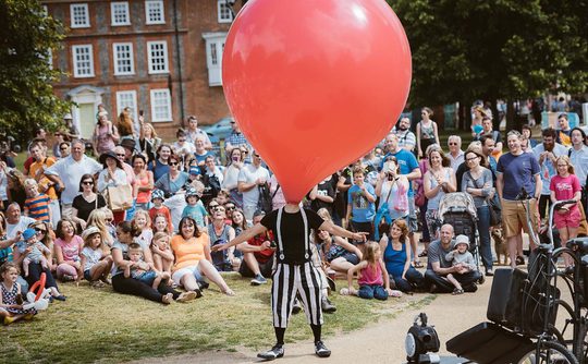 A outdoor performer wearing a big red balloon over his head, performing to a crowd.