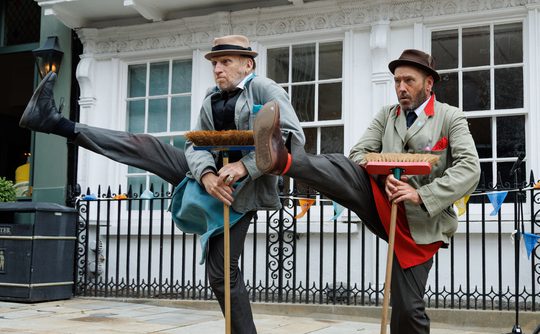 Two performers outdoors holding their legs in the air and balancing with brooms.