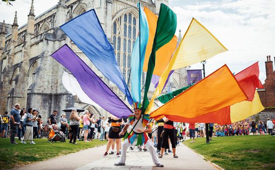 Performer standing in front of Winchester Cathedral with multi-coloured flags attached to their back.