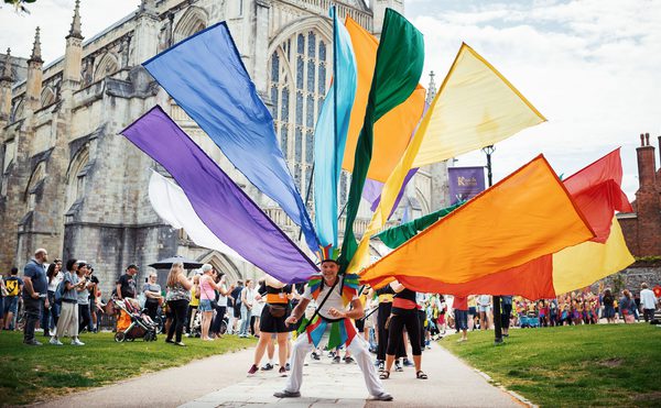 Performer standing in front of Winchester Cathedral with multi-coloured flags attached to their back.