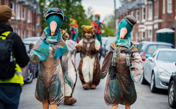 Performers dressed as ducks walking down a street with car parked either side.
