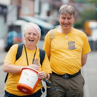 Two Hat Fair volunteers smile in a street while holding a fundraising bucket, both wearing yellow Hat Fair t-shirts.
