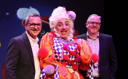 Two people in suits stood either side of a panto dame.