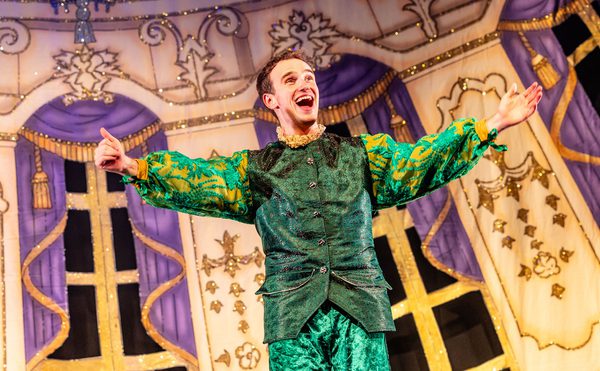 A panto performer dressed in shiny green clothing, looking happy, with their arms outstretched.