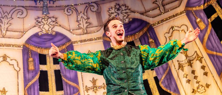 A panto performer dressed in shiny green clothing, looking happy, with their arms outstretched.