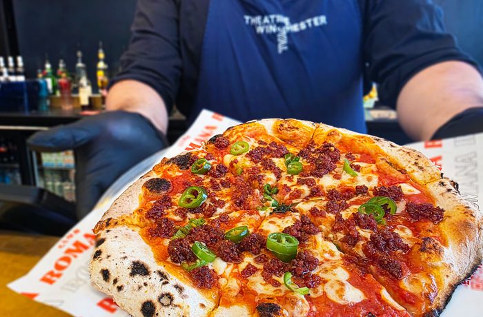 A person in a Theatre Royal Winchester apron holding out a cooked pizza.