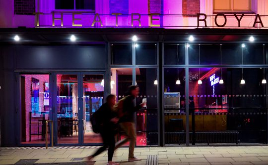 Two blurry people walking past Theatre Royal Winchester at night.
