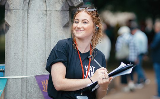 A person wearing a black Hat Fair t-shirt, smiling, and holding clipboard and pen.