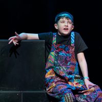 A young person sat on a box wearing dungarees and talking.
