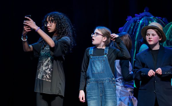 Young people performing on stage, one holding up a phone.