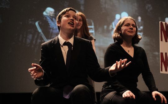 Young performers dressed smartly, sat on stage, one gesturing with their arms.