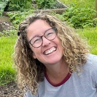 A person with a big smile, long curly brown hair and glasses crouch in their garden.