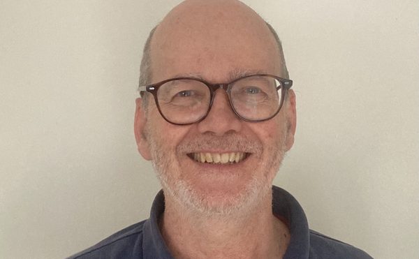 A person in a blue polo shirt and glasses stood in front of a plain white wall.