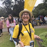 A person in a yellow Hat Fair t-shirt wearing a hat and glasses, and holding a fundraising bucket.