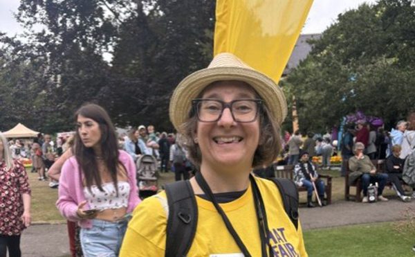 A person in a yellow Hat Fair t-shirt wearing a hat and glasses, and holding a fundraising bucket.