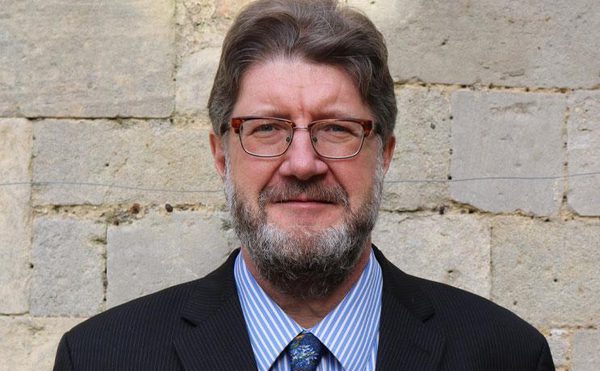 A person stood in front of a pale brick wall. They're wearing a dark blazer, shirt and tie. They have a beard and glasses.